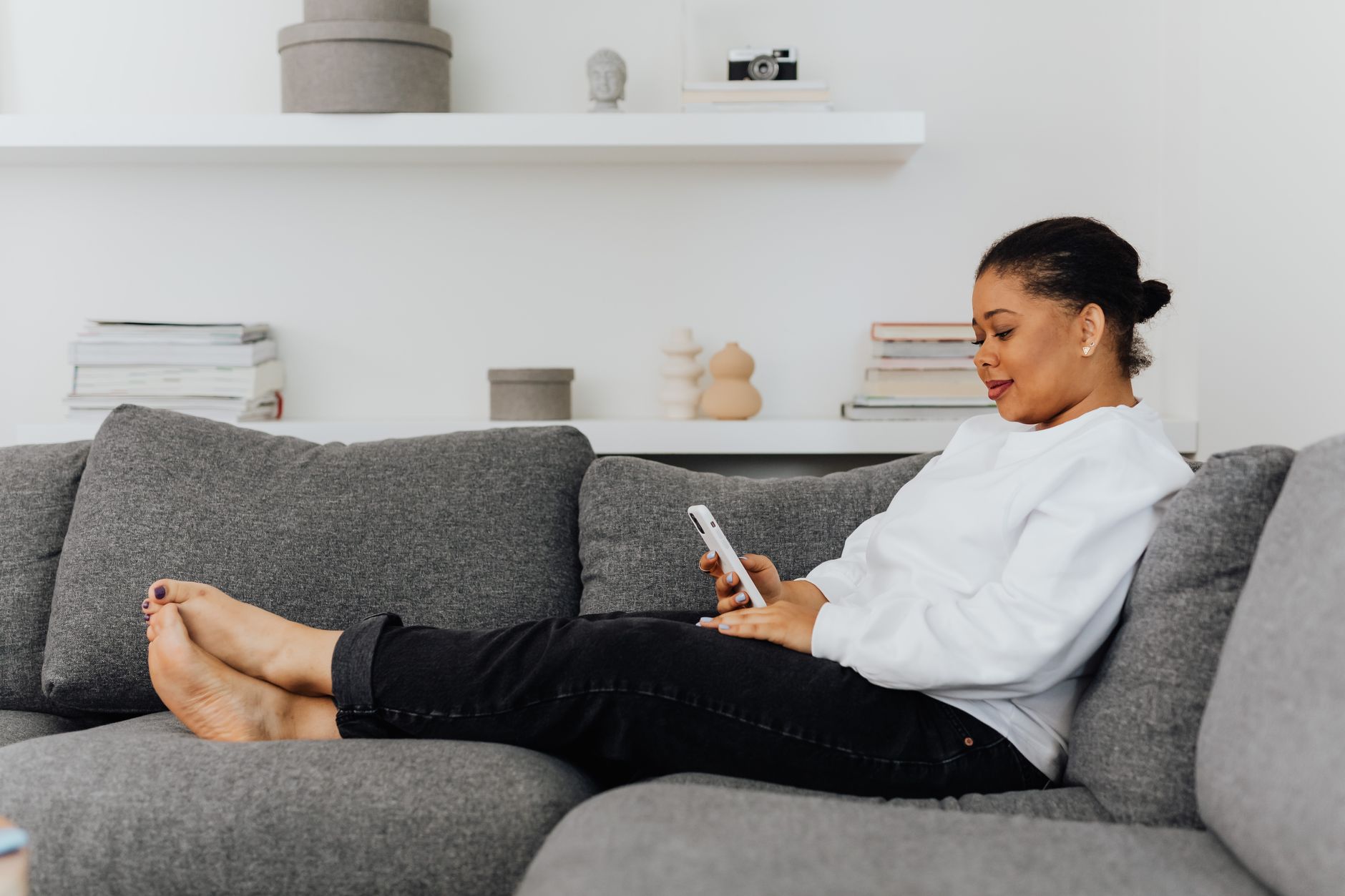 Una donna seduta comodamente su un divano grigio, in un soggiorno luminoso, sorride mentre guarda lo schermo dello smartphone che tiene in mano.